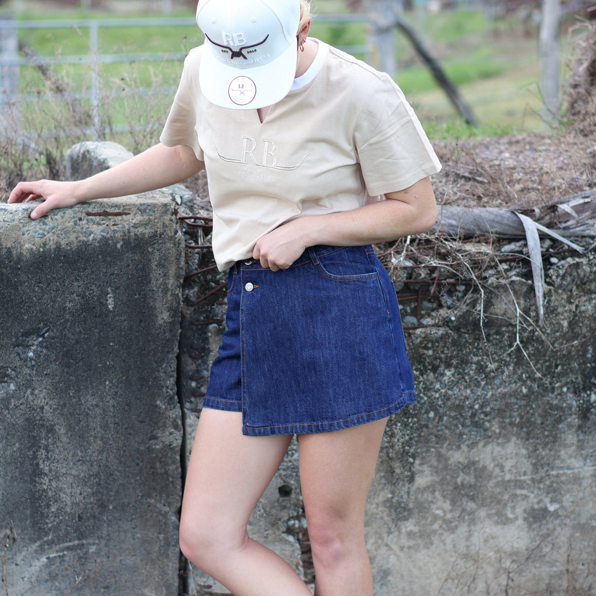 Model wearing an Angel Wings dark denim skort, beige Rare Breed tee and white cap beside a rustic outdoor wall.