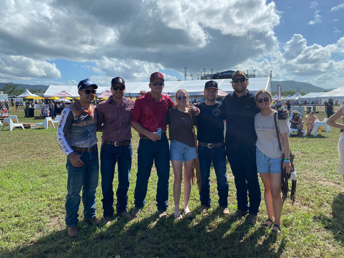 Seven friends standing together on grass at an outdoor festival, wearing shirts, tees, jeans, shorts and caps.